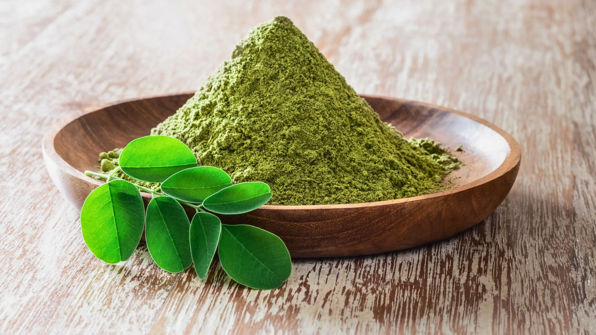Moringa powder heaped in a wooden bowl with fresh moringa leaves